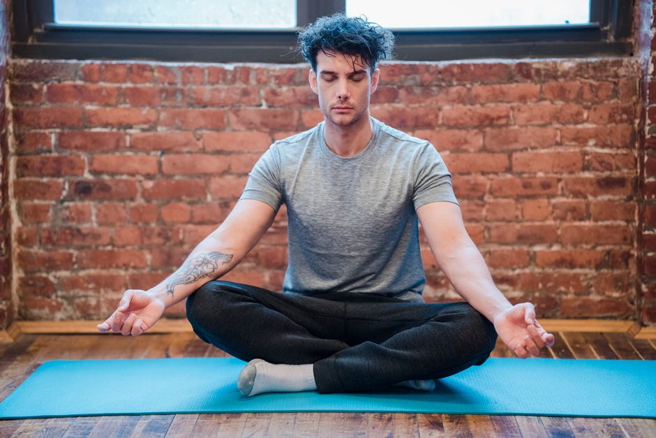 Full body of focused male with closed eyes doing Sukhasana while meditating on yoga mat against brick wall